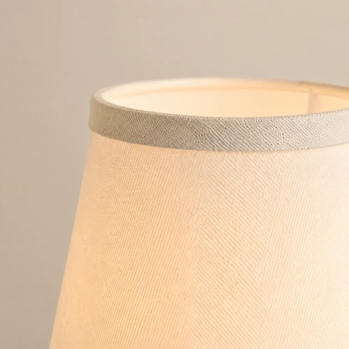 Close-up of a fabric lampshade in a warm cream or beige tone with a textured woven band around the top edge, shown against a plain light gray background.