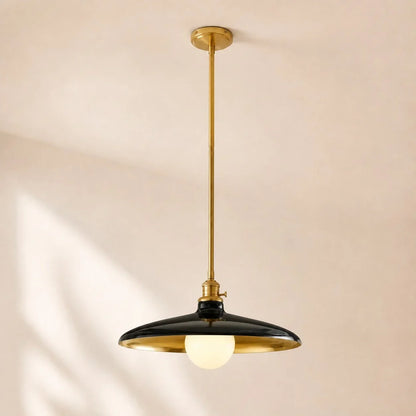 A modern pendant light fixture with a black dome-shaped shade, gold-toned metal frame and canopy, and a glowing warm white globe bulb, suspended from a ceiling against a warm beige wall.