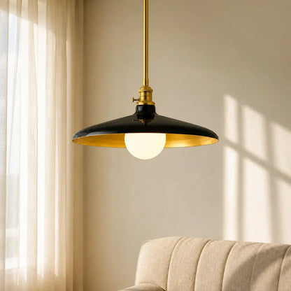 Pendant light with black shade and gold trim hanging in a room with beige curtains and sofa.
