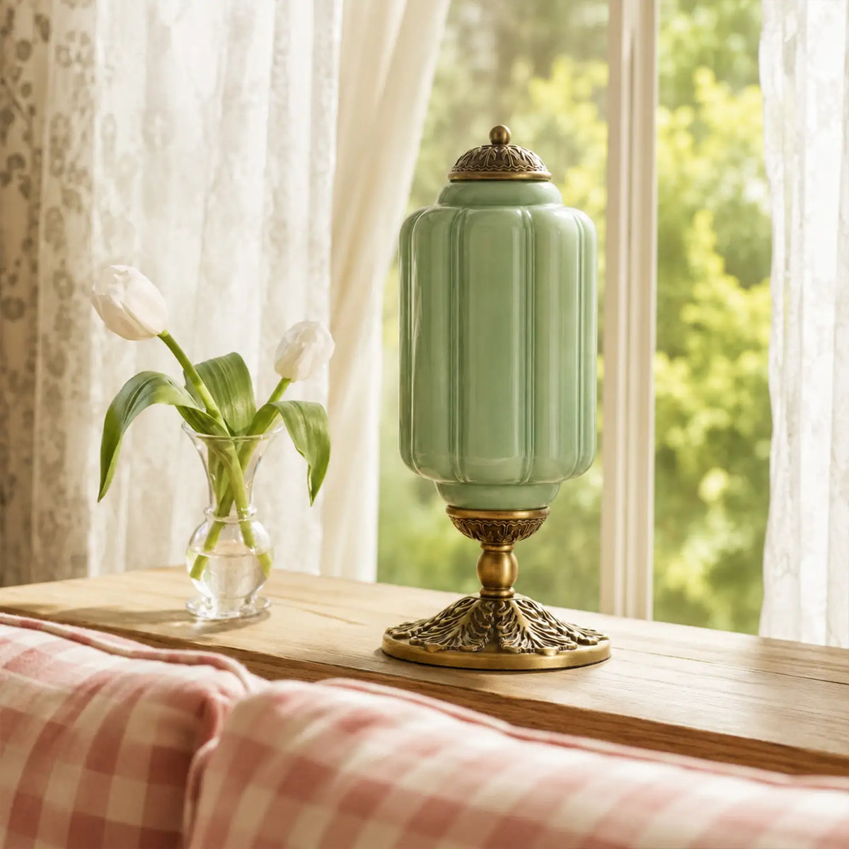 Decorative green lamp with gold base on a wooden surface near a window with white curtains.