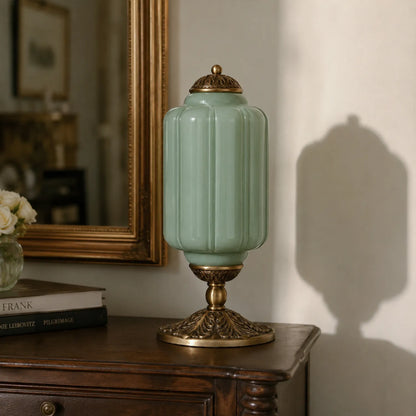 Decorative green lamp with gold base on a wooden surface, with books and a mirror in the background.
