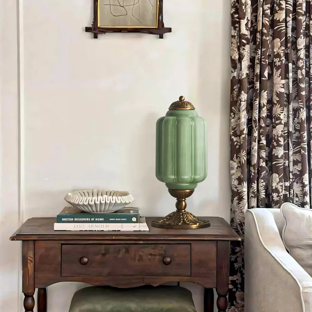 Green lamp on a wooden side table with books and a decorative bowl, next to a floral curtain.